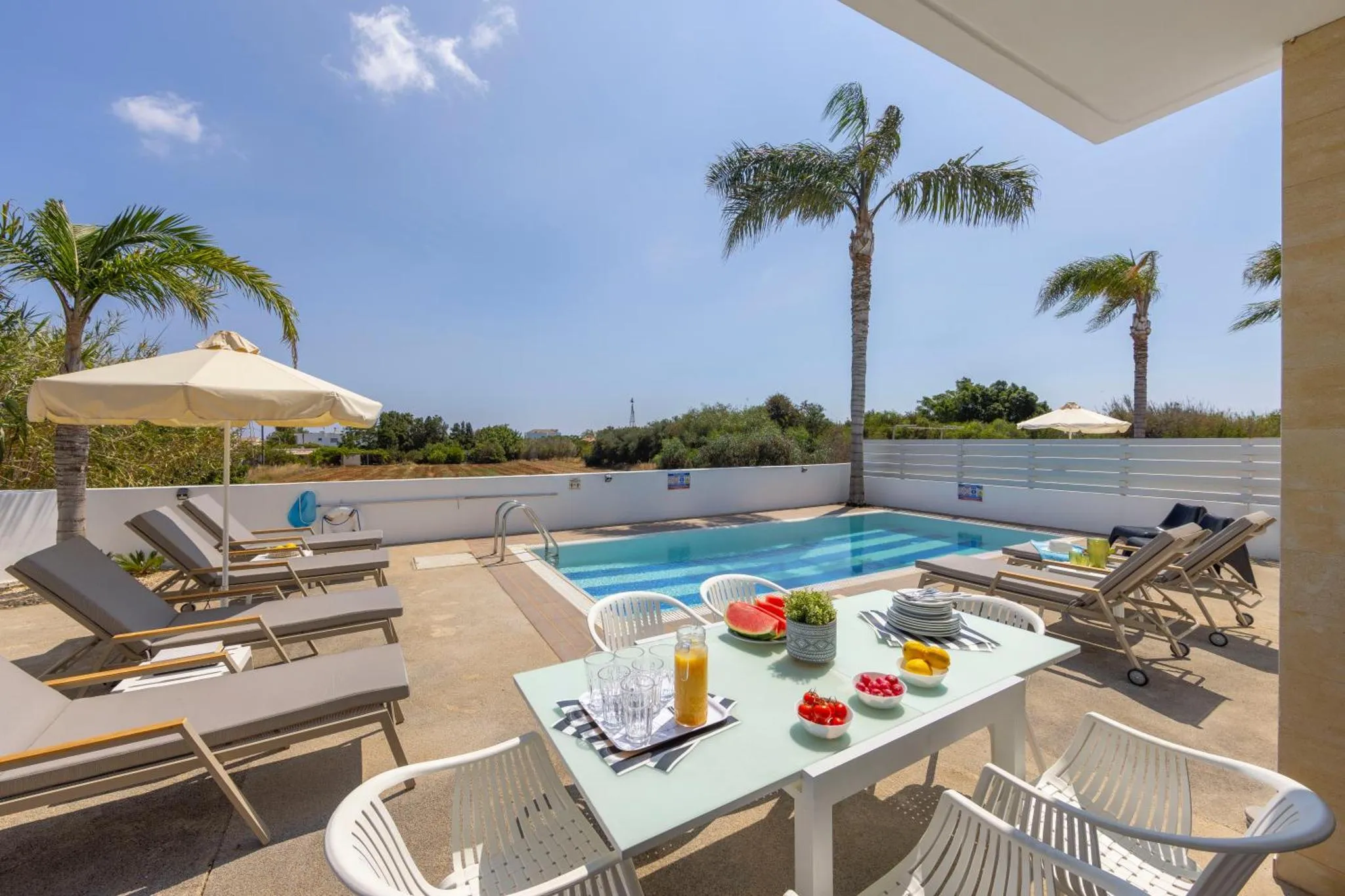 Une table de repas est mise en scène au bord d'une piscine avec des transats et des palmiers.