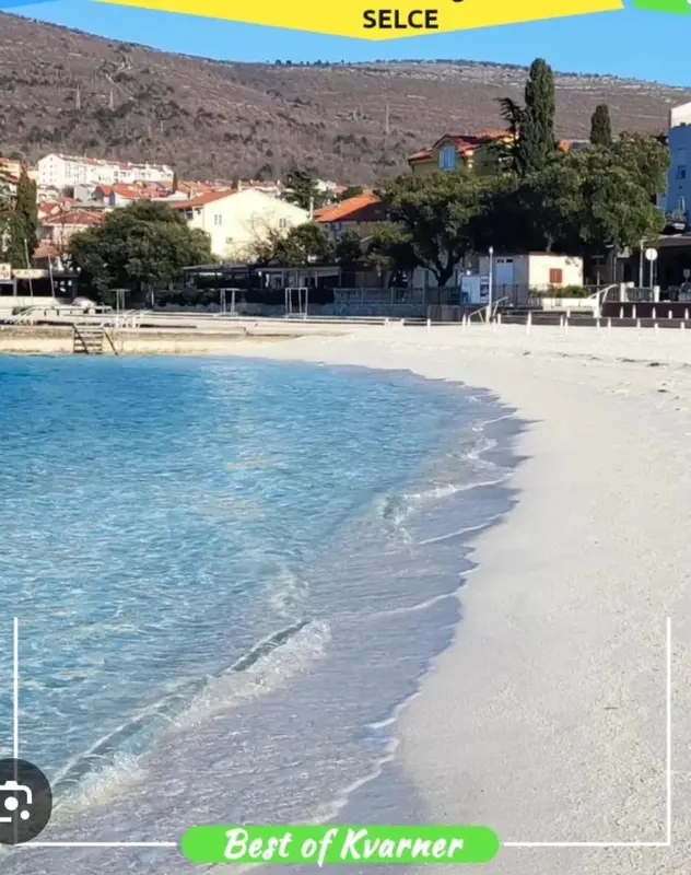 Une plage de galets bordée d'eau turquoise avec une vue sur des bâtiments et une végétation en arrière-plan.