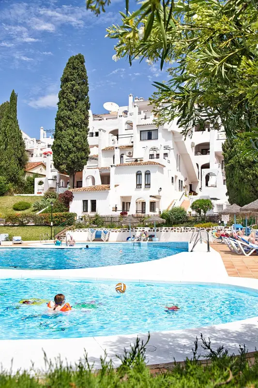Bâtiment blanc à plusieurs étages avec piscine au premier plan et végétation luxuriante.