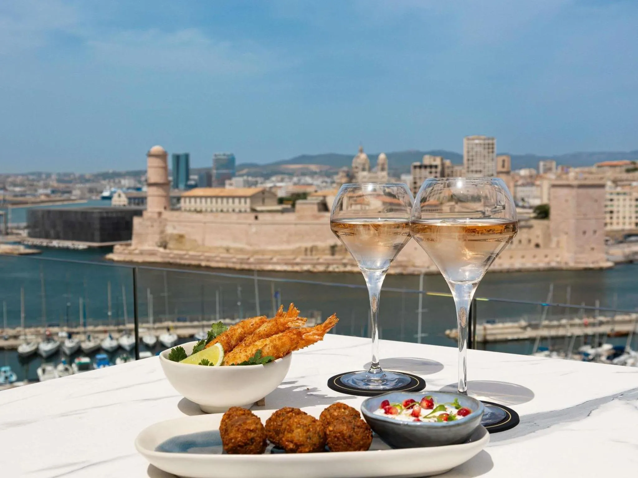 Deux verres de vin rosé et une assiette de tapas sont posés sur une table en marbre blanc avec vue sur la mer et la ville.