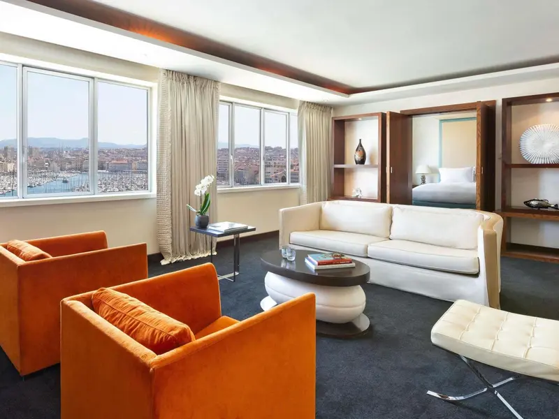 Un salon moderne avec canapés et fauteuils face à une grande fenêtre offrant une vue sur la ville et le port.