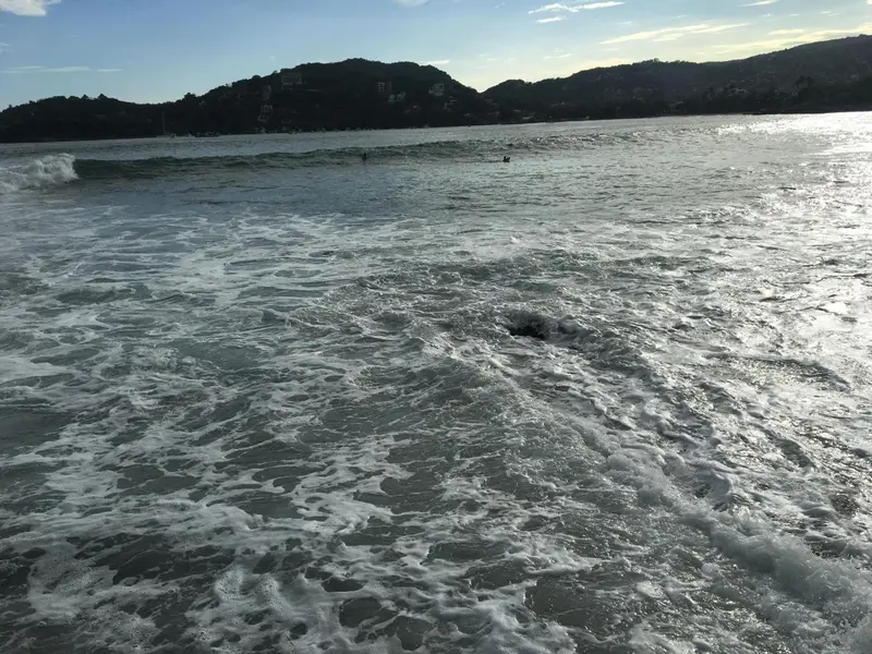 Vagues déferlant sur une plage de sable fin avec une côte montagneuse en arrière-plan.