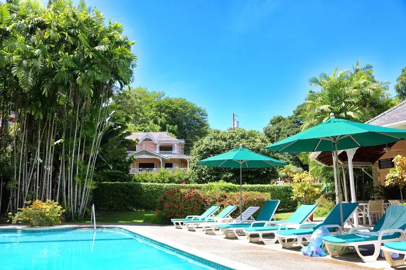 Transats bleus alignés au bord d'une piscine turquoise, sous des parasols verts, dans un jardin tropical.
