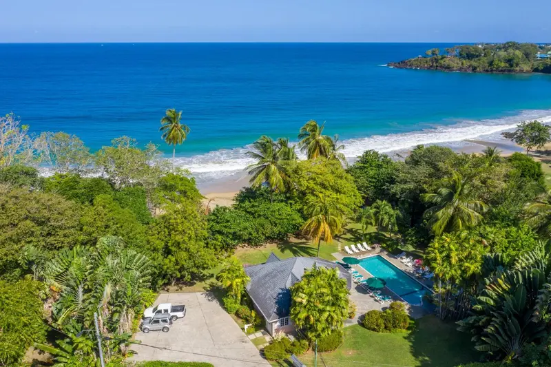 Vue aérienne d'une plage bordée de végétation luxuriante et d'une piscine près de bâtiments.