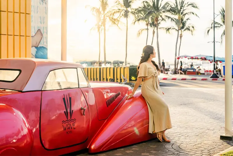 Une femme se tient près d'une voiture rouge vintage sur une promenade en bord de mer.