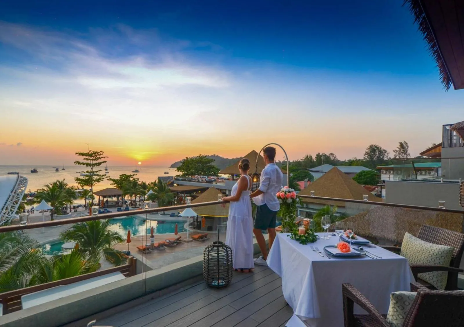 Un couple admire le coucher de soleil depuis une terrasse avec une table dressée pour un dîner romantique.