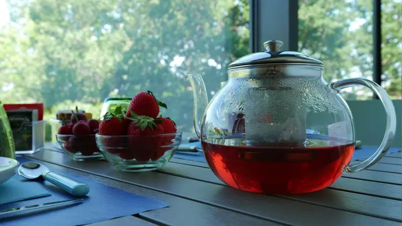 Une théière en verre pleine de boisson rouge et des fraises fraîches sur une table.