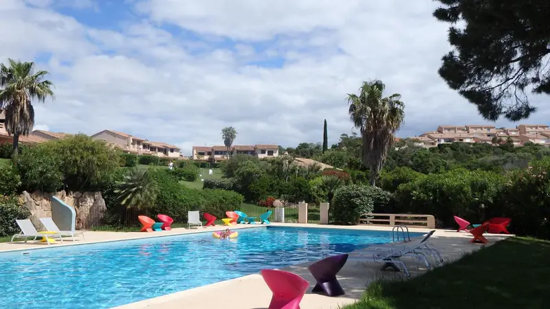 Une piscine turquoise bordée de chaises longues colorées, entourée de végétation luxuriante et de bâtiments.