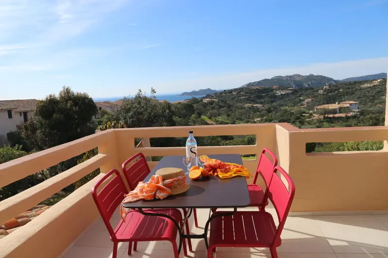 Une table avec des couverts est dressée sur une terrasse ensoleillée avec vue sur la mer et les collines.