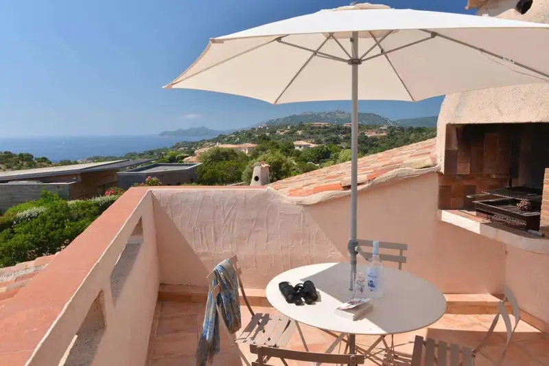 Une terrasse avec une table ronde et un parasol offre une vue sur la mer et les collines.