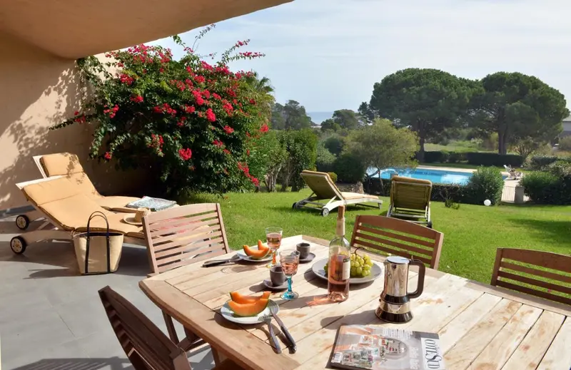 Table de repas en extérieur avec boissons et fruits, devant une pelouse verdoyante et des transats.