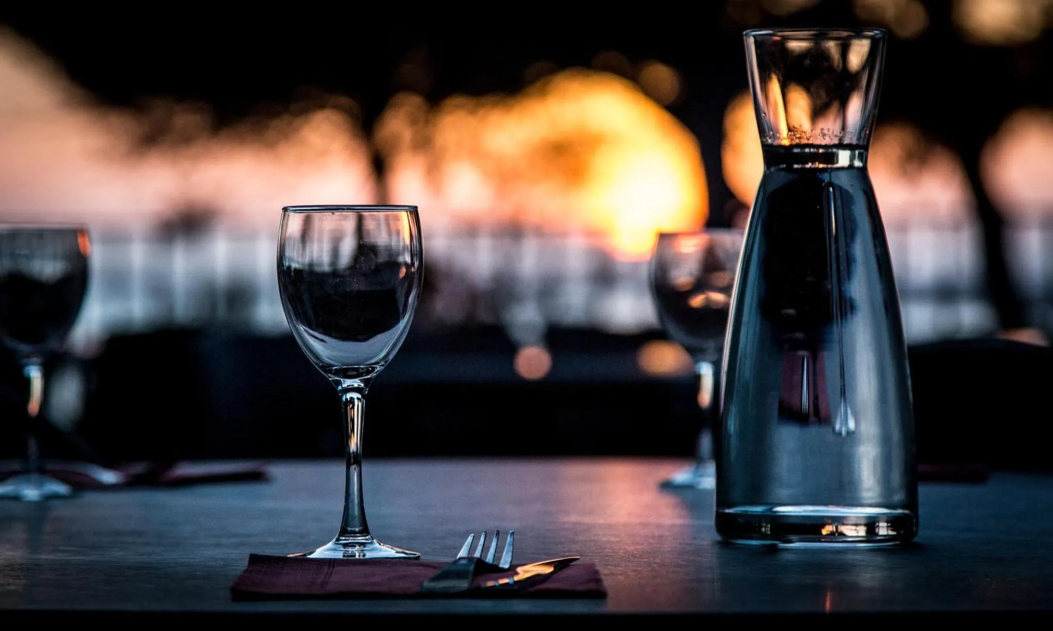 Une table est dressée avec des verres et des couverts, face à un coucher de soleil sur l'eau.