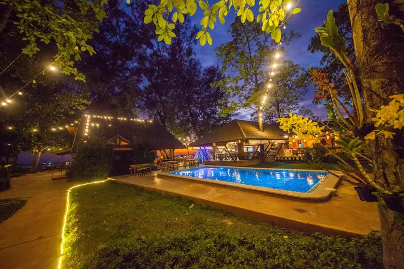 Piscine illuminée au crépuscule avec végétation luxuriante et bâtiment en arrière-plan.