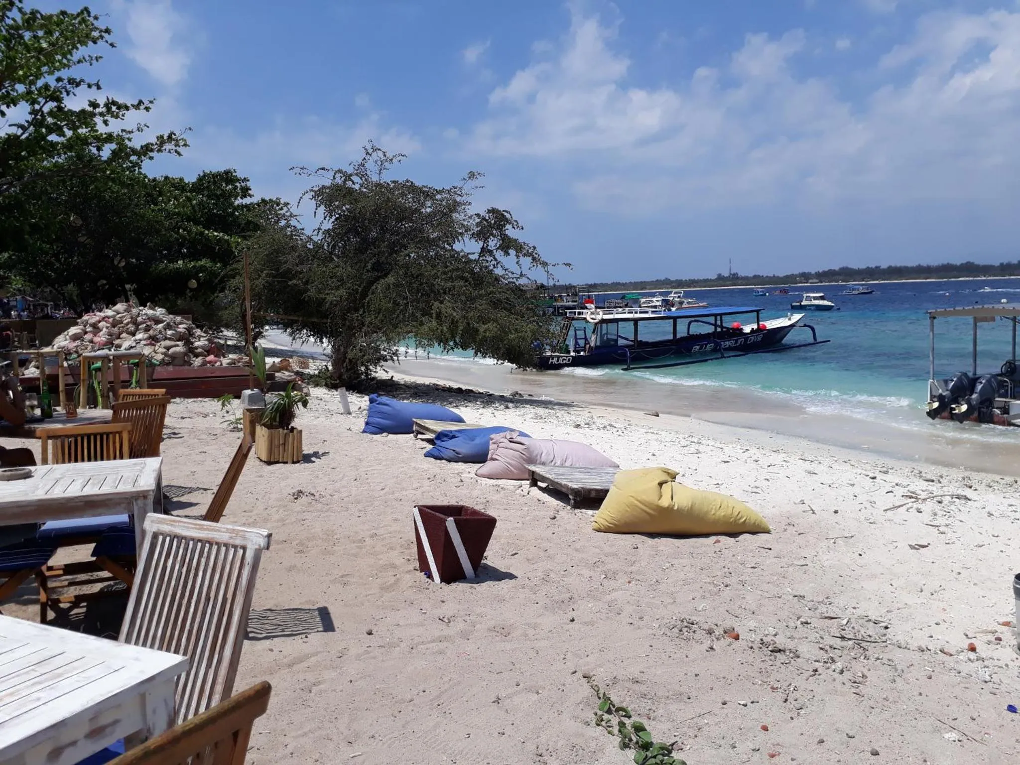 Une plage de sable blanc avec des transats et des tables, face à la mer turquoise et des bateaux.
