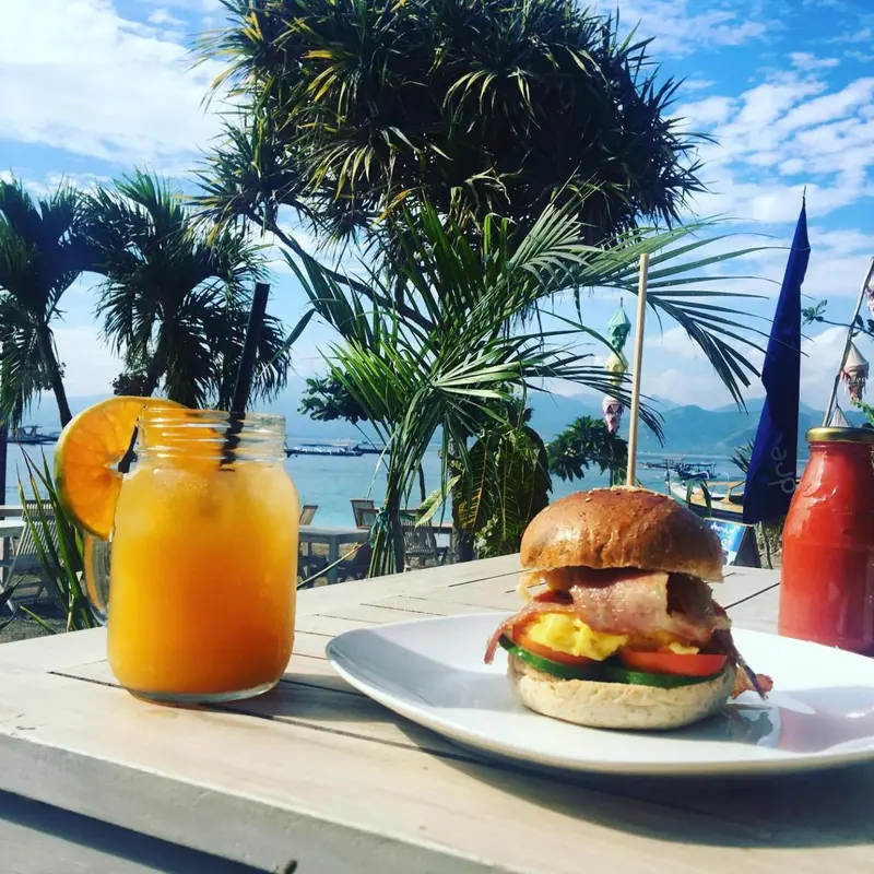 Une table en bois présente des boissons et un hamburger en bord de mer.
