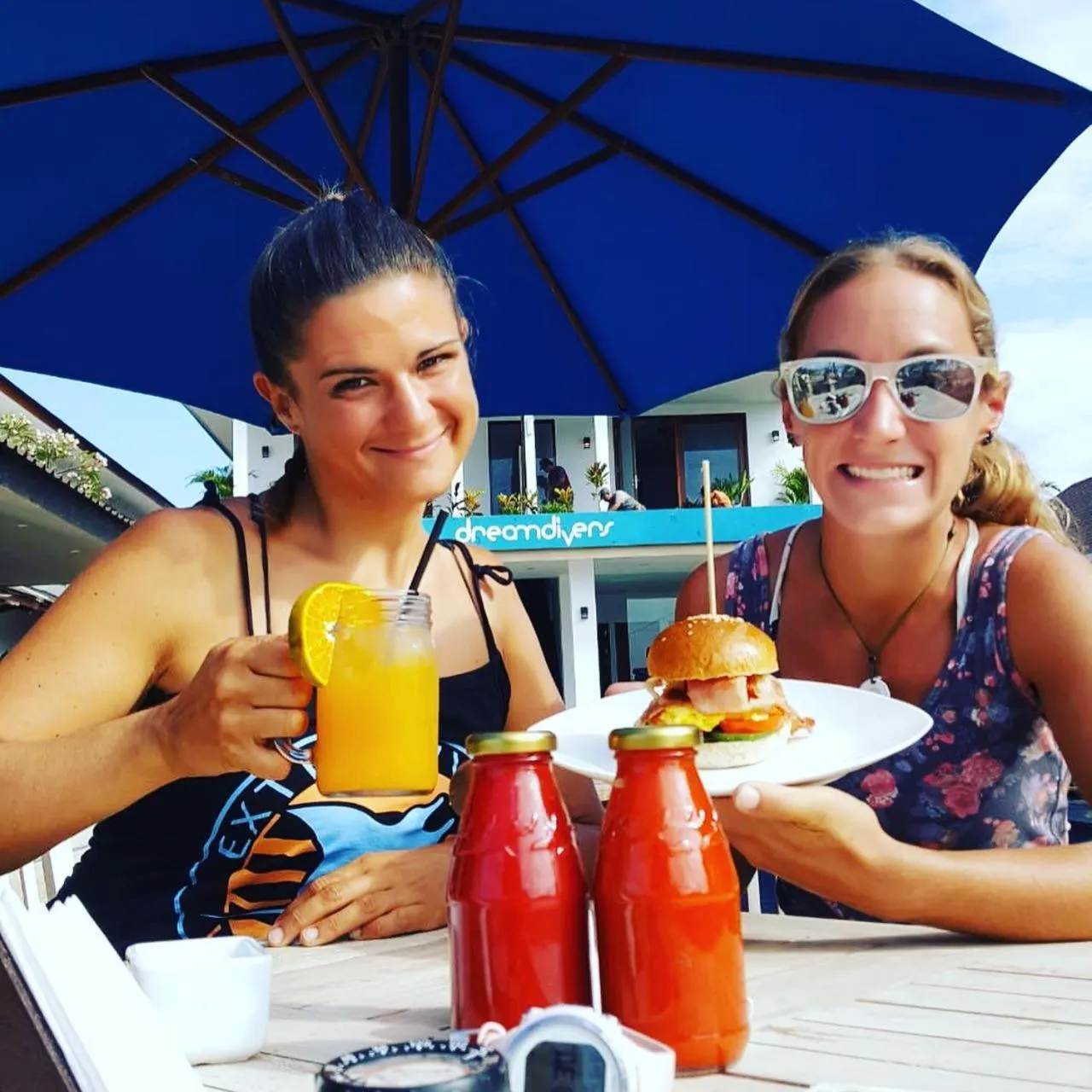 Deux femmes souriantes déjeunent en terrasse avec des boissons et des hamburgers.