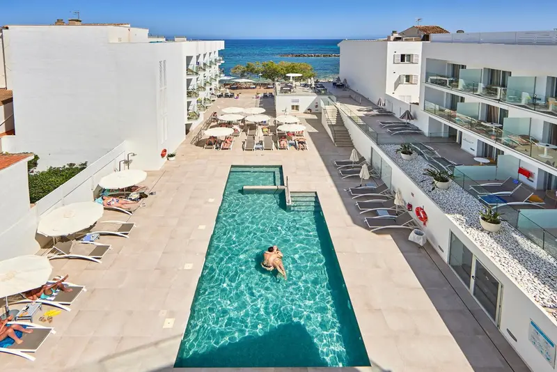 Vue aérienne d'une piscine rectangulaire entourée de transats et de parasols, avec des bâtiments blancs en arrière-plan.