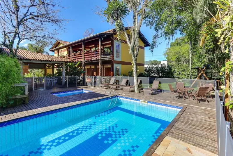 Une piscine rectangulaire turquoise est entourée d'une terrasse en bois avec des chaises longues et un bâtiment jaune et orange à l'arrière-plan.