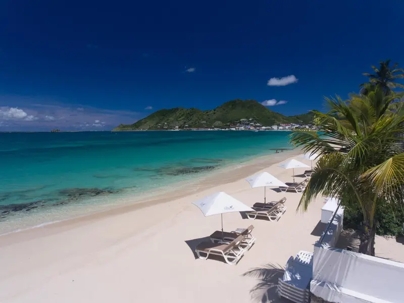 Plage de sable blanc bordée de transats sous des parasols, avec une mer turquoise et une colline verdoyante en arrière-plan.