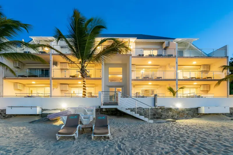 Bâtiment blanc éclairé avec des balcons et des transats sur une plage de sable.