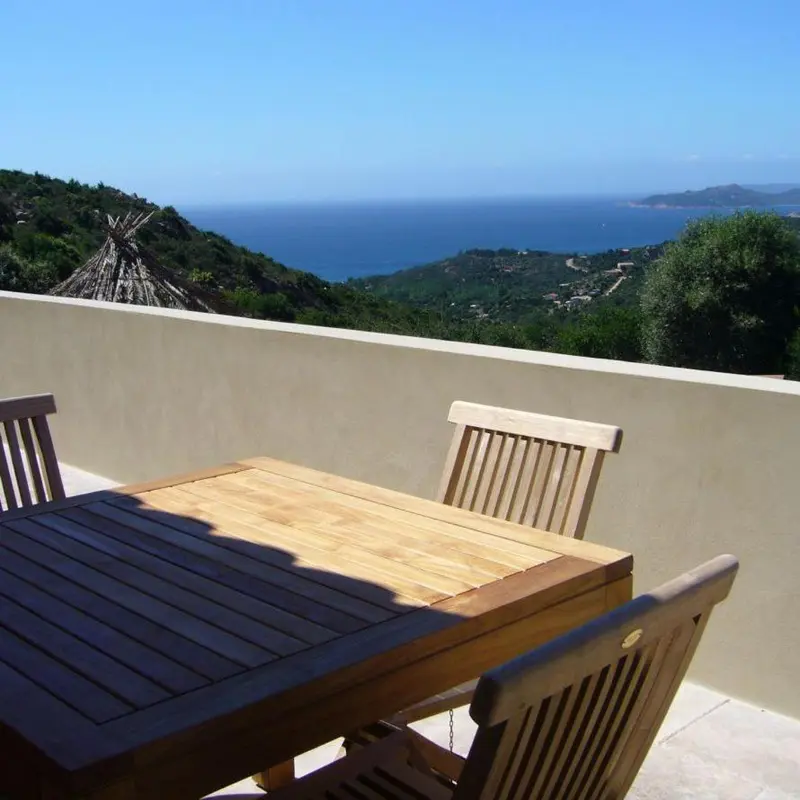 Une table en bois sur une terrasse ensoleillée avec vue sur la mer et les collines.