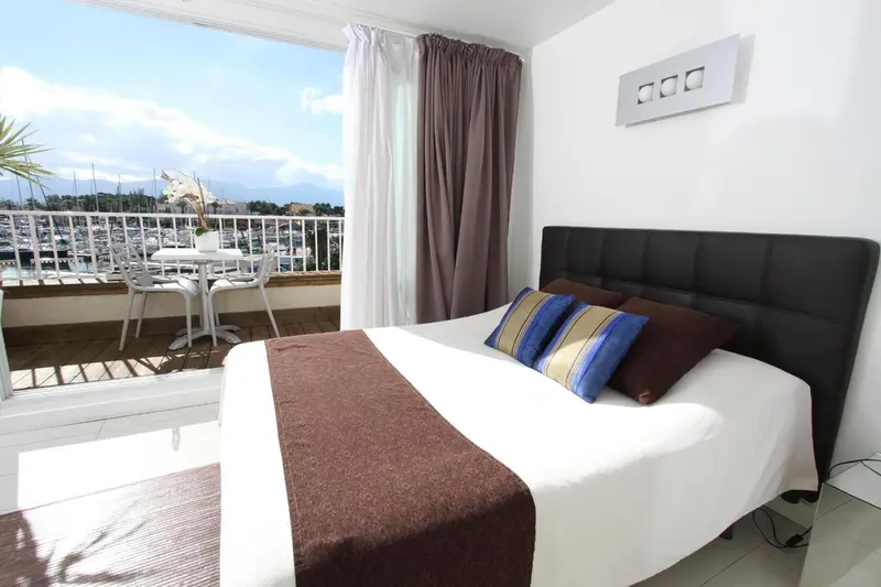 Une chambre lumineuse s'ouvre sur une terrasse avec vue sur un port de plaisance.