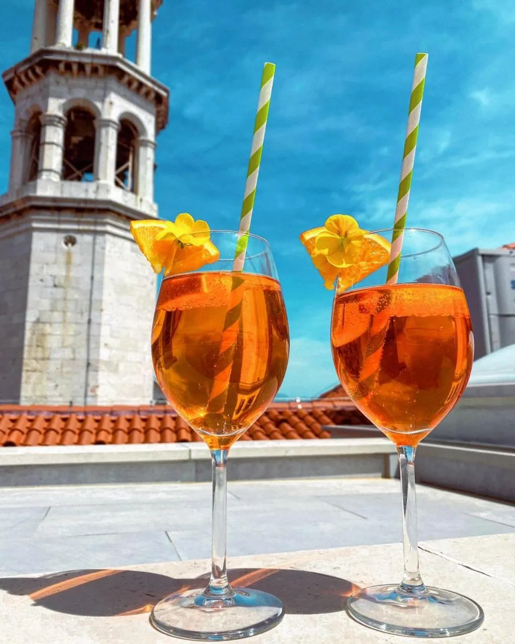 Deux verres de cocktail orange garnis de fleurs sur une terrasse ensoleillée avec une vue sur un bâtiment historique.