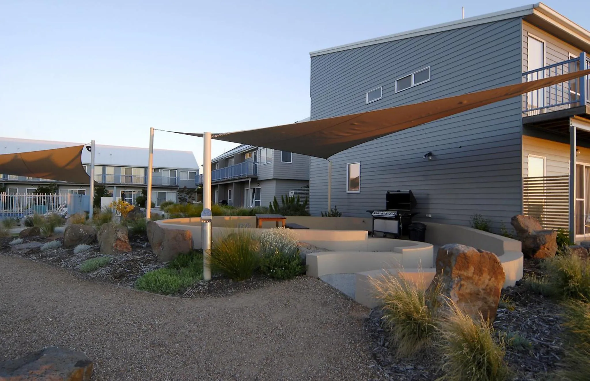 Terrasse ombragée avec barbecue et mobilier de jardin devant des bâtiments modernes.