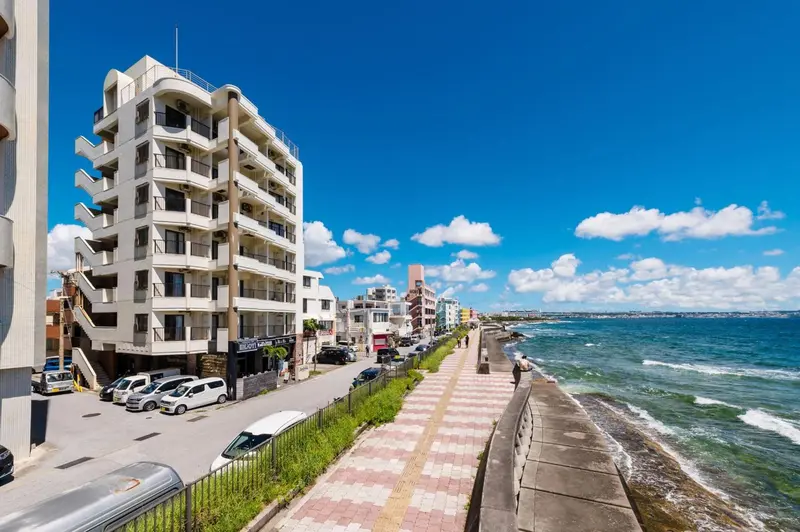 Bâtiment blanc à plusieurs étages avec des balcons, le long d'une promenade côtière ensoleillée.