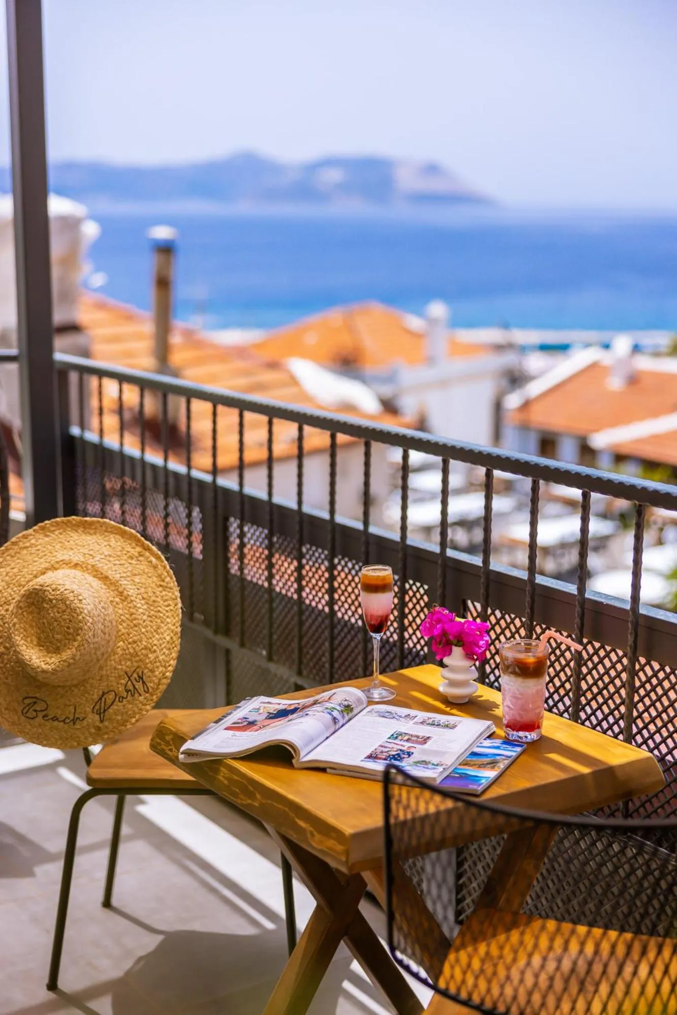 Une table avec un chapeau de paille, des magazines et des boissons sur un balcon avec vue sur la mer.
