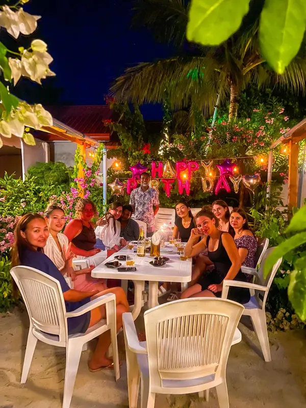 Une table conviviale est entourée de personnes assises sur des chaises blanches, dans un cadre tropical illuminé.