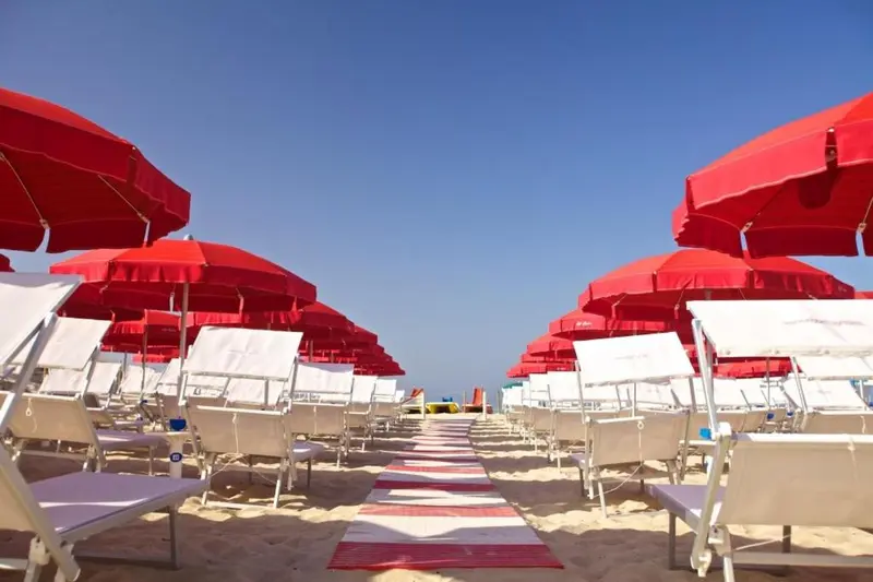 Transats blancs alignés sous des parasols rouges sur une plage de sable.