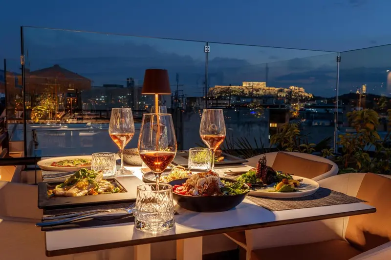 Une table dressée avec des plats et des boissons, face à une vue nocturne sur la mer et la ville.