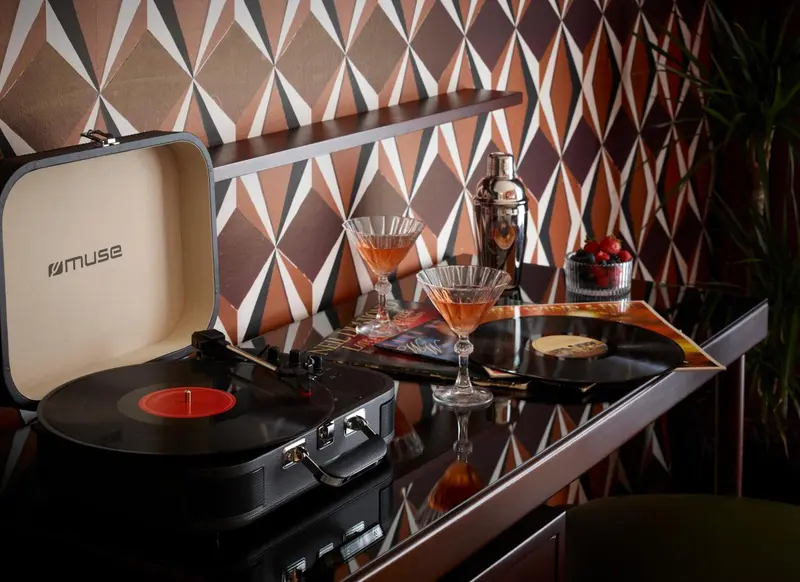 Une platine vinyle et des cocktails sont posés sur une table brillante devant un mur à motifs géométriques.