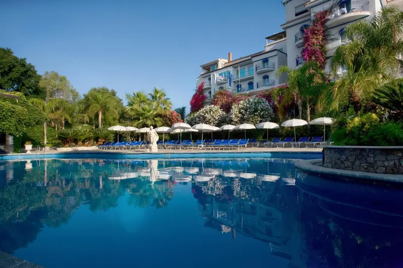 Piscine bleue entourée de transats et de végétation luxuriante devant un bâtiment blanc.