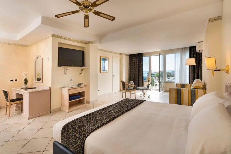 Chambre lumineuse avec un grand lit et un espace salon ouvert sur une vue extérieure.