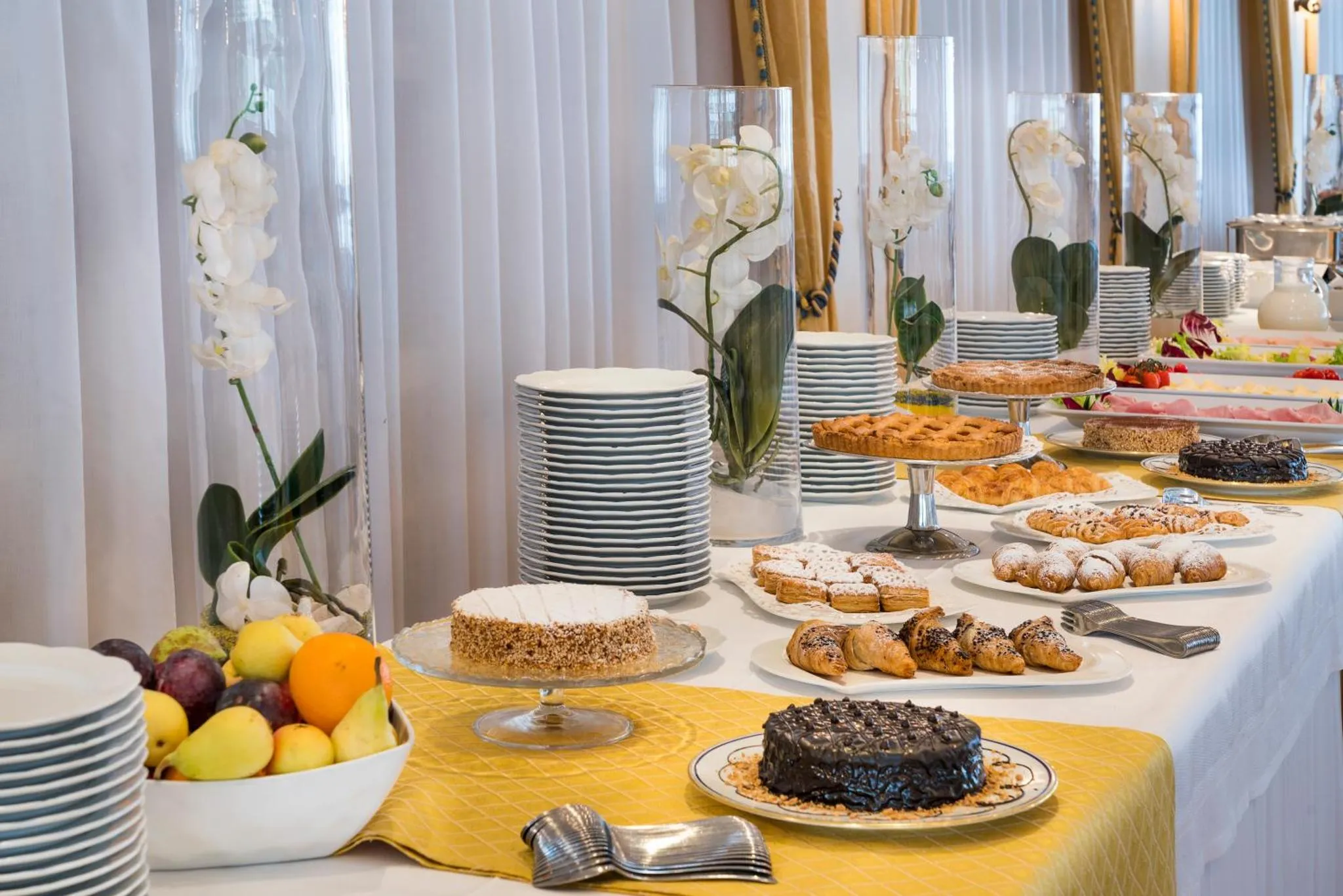 Une table garnie de pâtisseries, de fruits et de viennoiseries est présentée sur une nappe jaune.