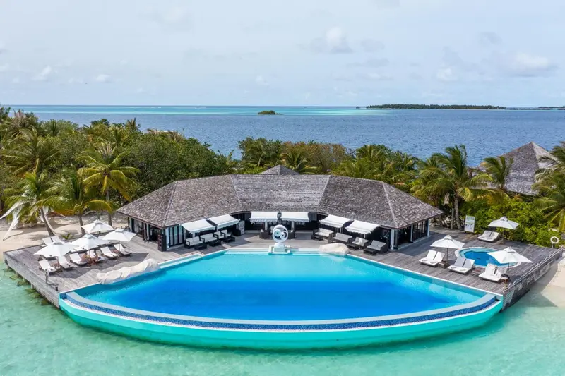 Vue aérienne d'une piscine turquoise bordée de transats blancs et d'un bâtiment à toit de chaume.