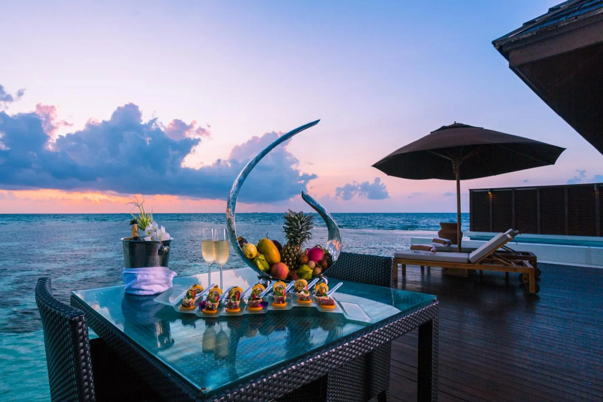 Une table dressée avec des mets et des boissons sur une terrasse en bois face à la mer au coucher du soleil.