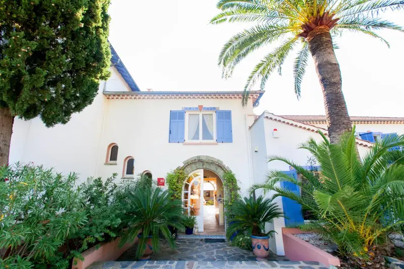 Bâtiment blanc à deux étages avec des volets bleus et une végétation luxuriante.