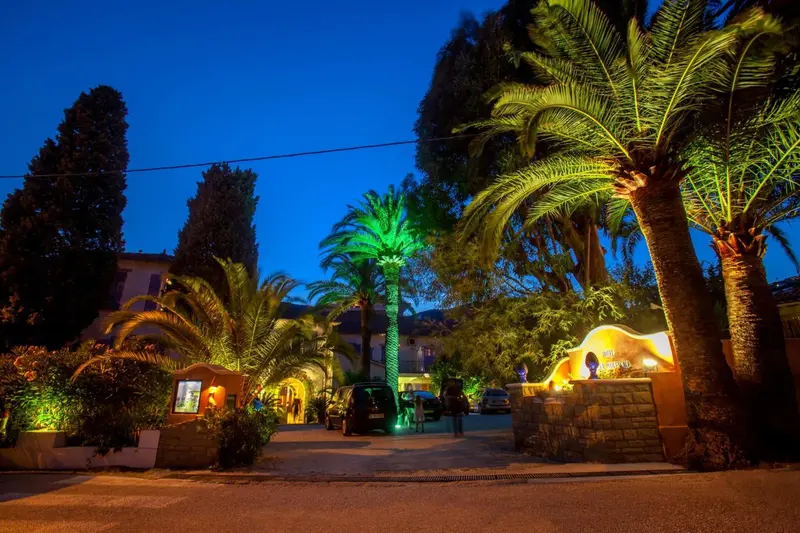 Entree d'un etablissement illuminé avec des palmiers et une végétation luxuriante.