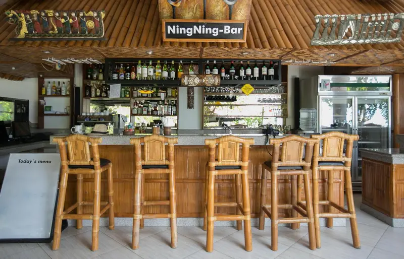 Un comptoir de bar en bois avec des tabourets hauts, sous un toit de chaume.