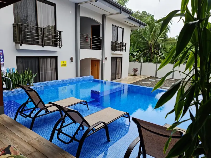 Transats bleus au bord d'une piscine turquoise devant un bâtiment blanc.