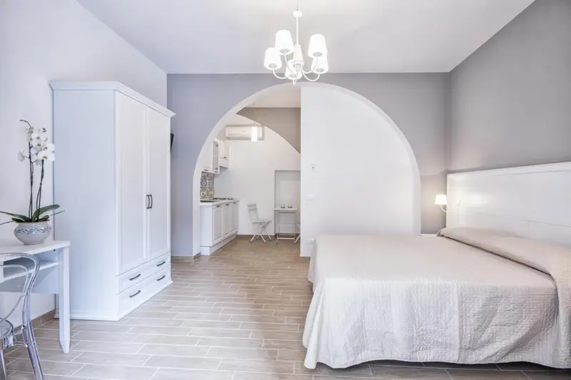 Une chambre lumineuse avec un lit recouvert de blanc et un espace cuisine.
