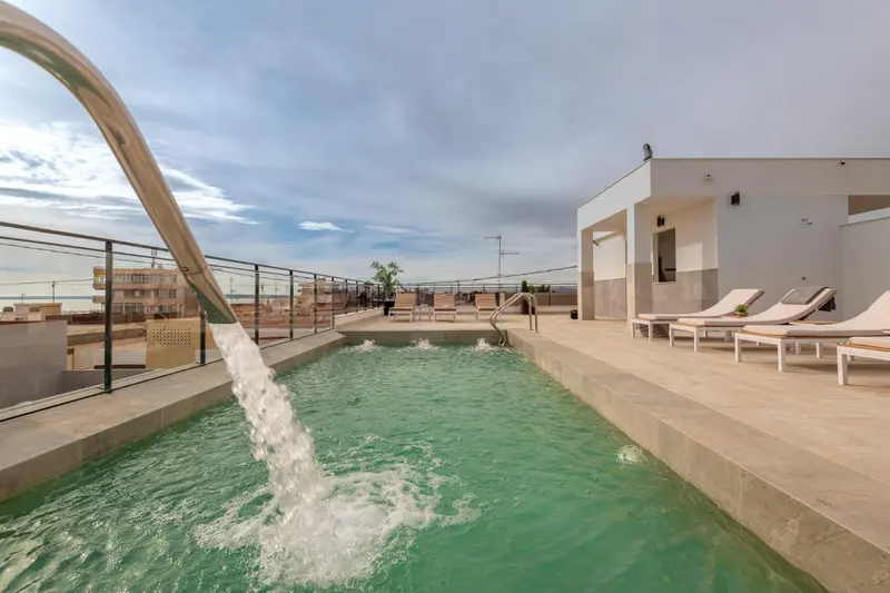Une cascade d'eau se déverse dans une piscine rectangulaire entourée d'un sol carrelé et de chaises longues.