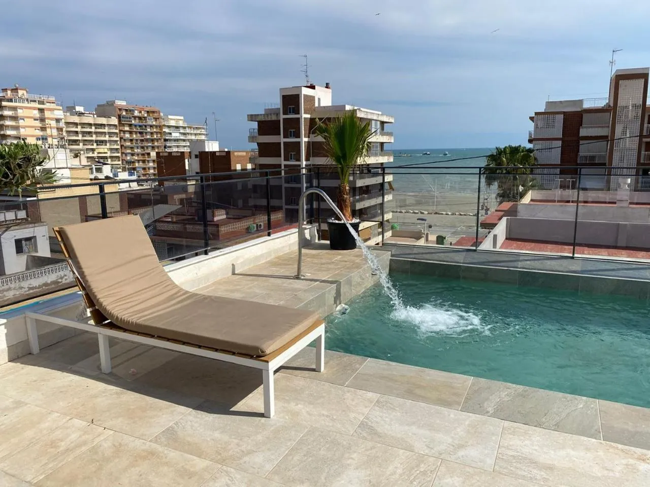 Transats et piscine sur une terrasse ensoleillée avec vue sur la mer et la ville.