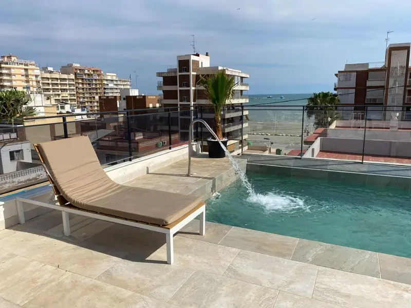 Transats et piscine sur une terrasse ensoleillée avec vue sur la mer et la ville.