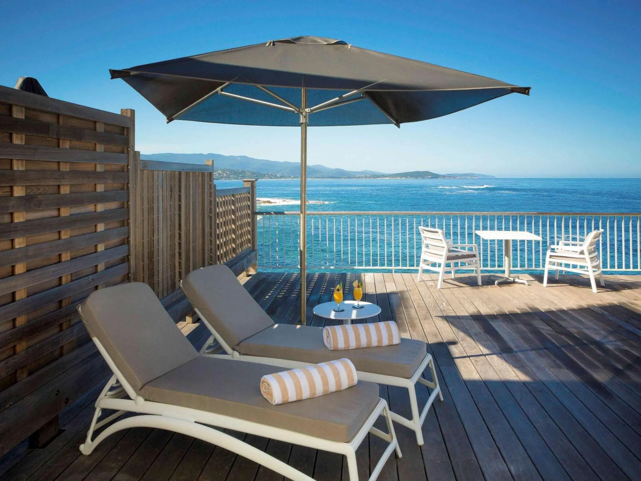 Deux transats et une table basse avec des boissons sur une terrasse en bois avec vue sur la mer.