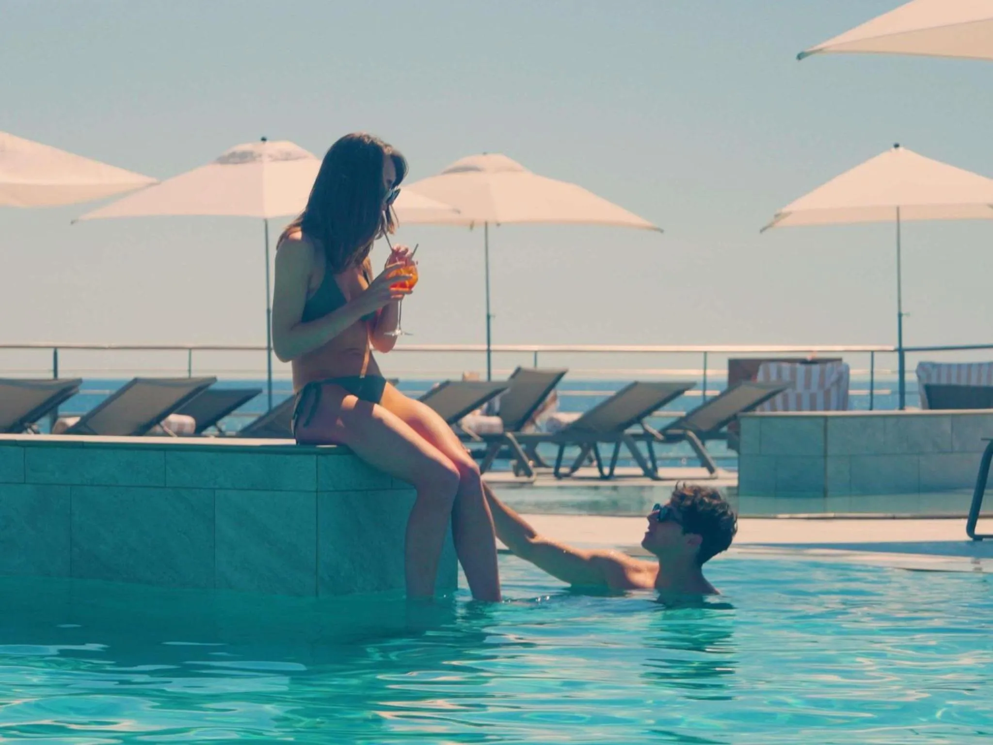 Une femme assise au bord d'une piscine turquoise déguste une boisson, tandis qu'un homme se détend dans l'eau.