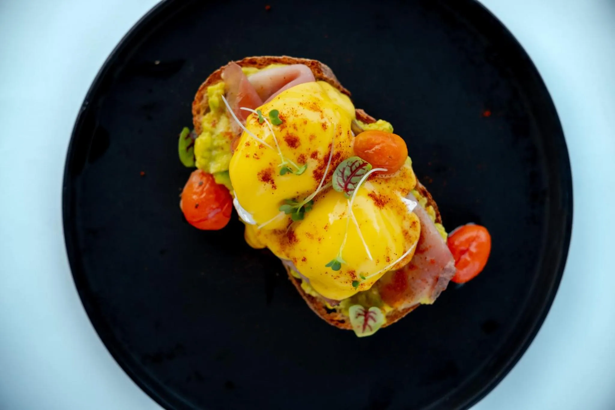 Une tartine généreusement garnie d'avocat, de jambon, d'oeufs pochés et de tomates cerises est présentée sur une assiette noire.